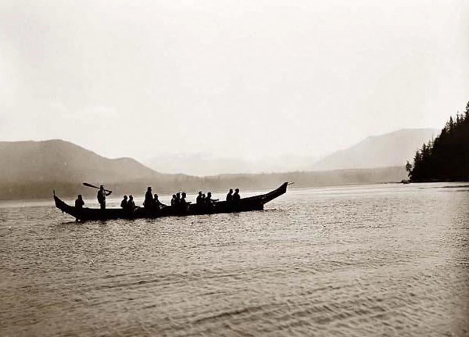 A large Northwest Coast canoe of paddlers silhouetted on misty water.