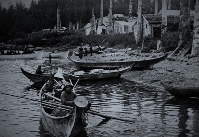 Men in dugout canoes on the water before a Northwest Coast village of plank houses and totem poles, more canoes drawn up on the beach.