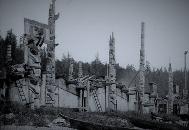 A row of Northwest Coast plank houses fronted by tall carved totem poles, canoes resting in the grass below.