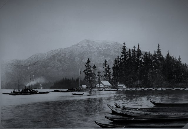 A line of canoes on a mountain-ringed inlet, with a steamer and a small settlement across the water.