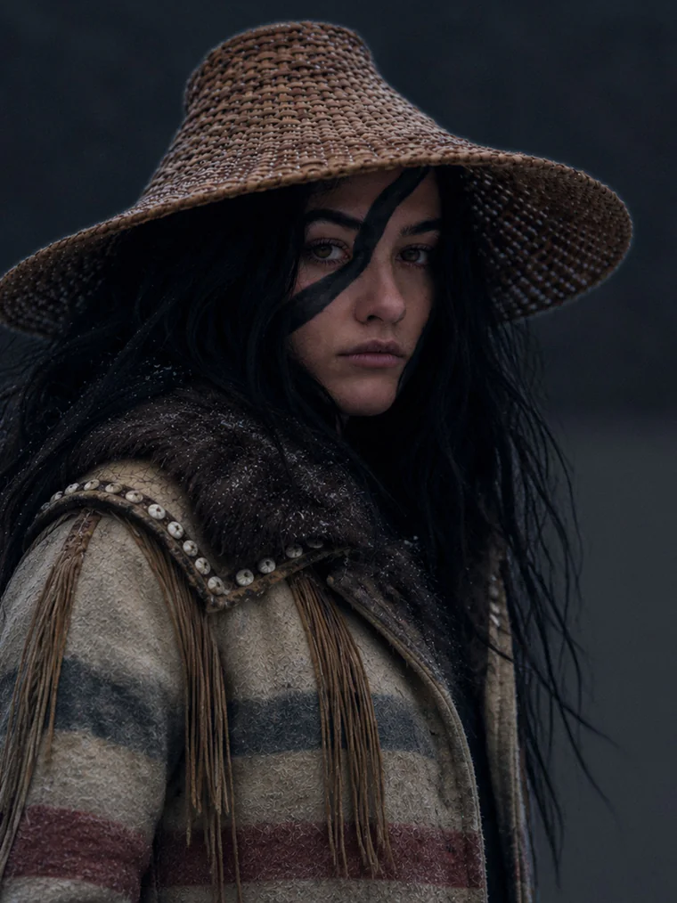 A young woman in a woven cedar hat and striped Hudson's Bay trade coat, face painted with a black formline stripe, standing in falling snow