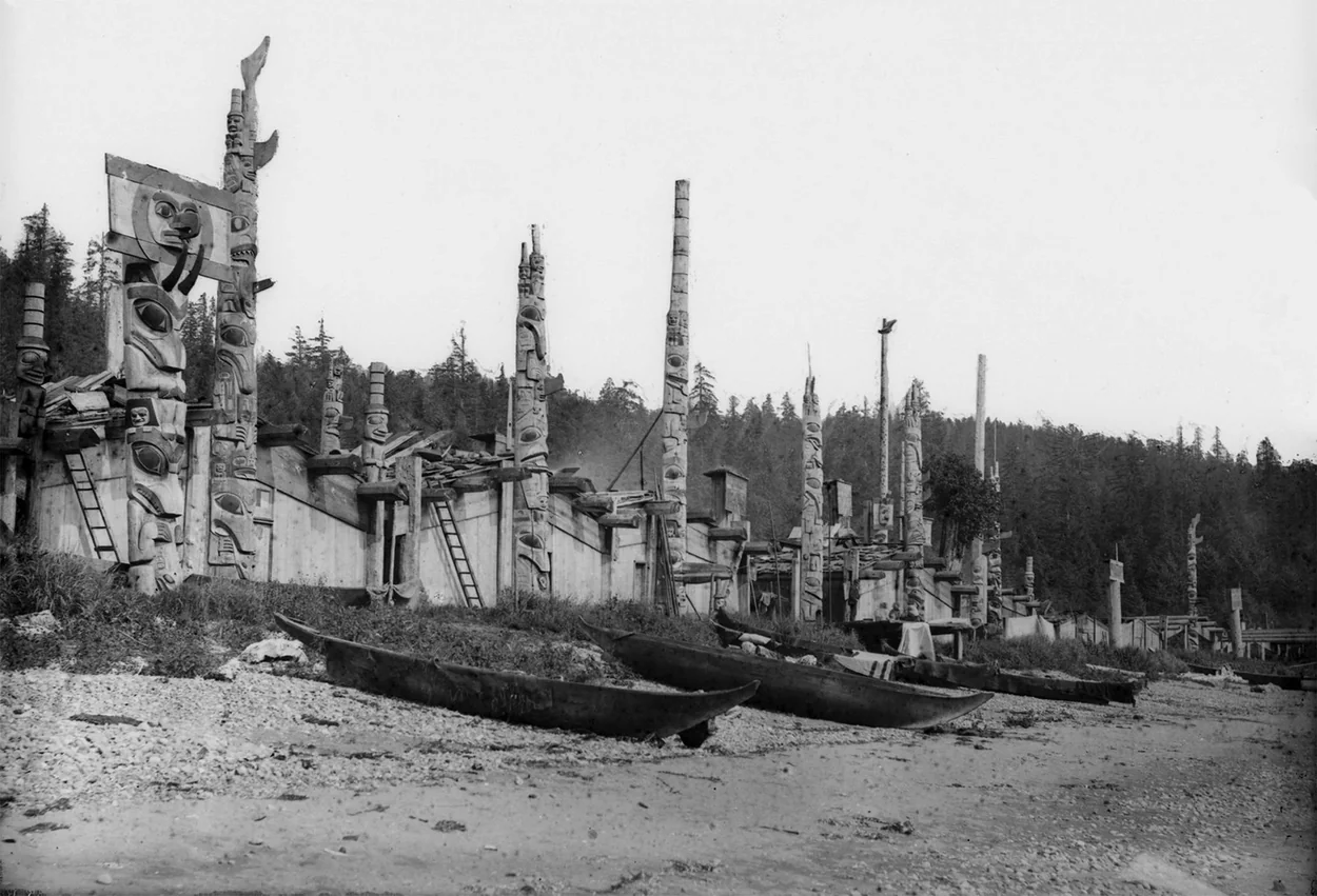 A Northwest Coast village of houses, totem poles, and canoes drawn up on the beach.